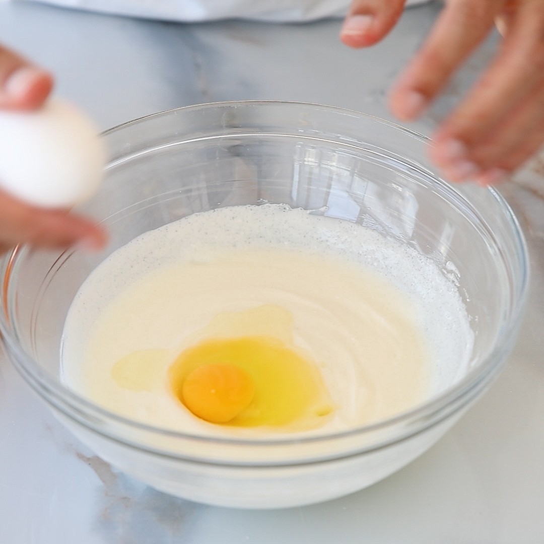 A person cracks an egg into a clear glass bowl with a creamy mixture on a marble countertop. Another hand holds an egg, ready to add it to the bowl.
