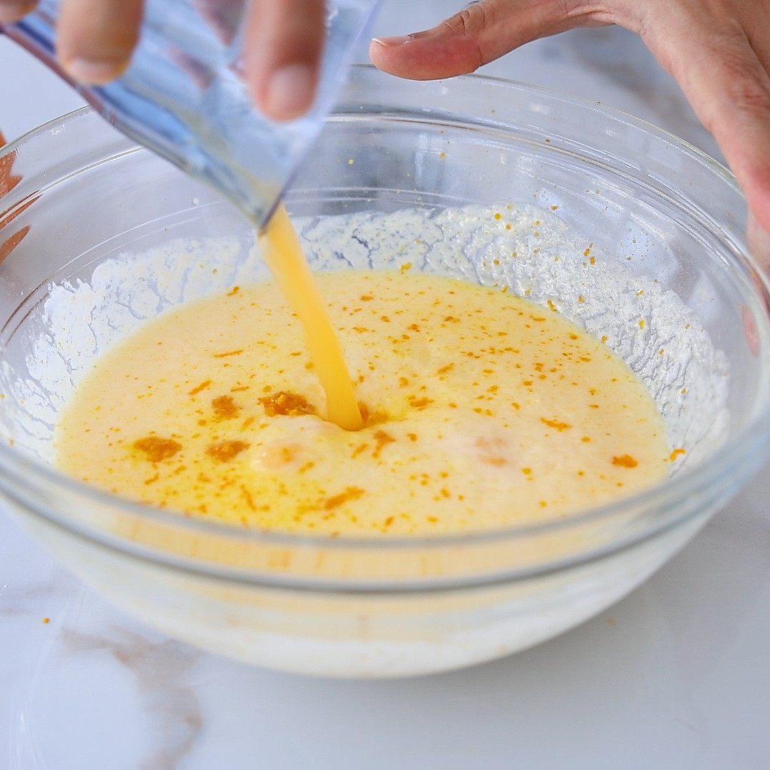 A hand pours orange juice from a glass into a large bowl with a creamy yellow mixture, likely for baking, on a white surface.