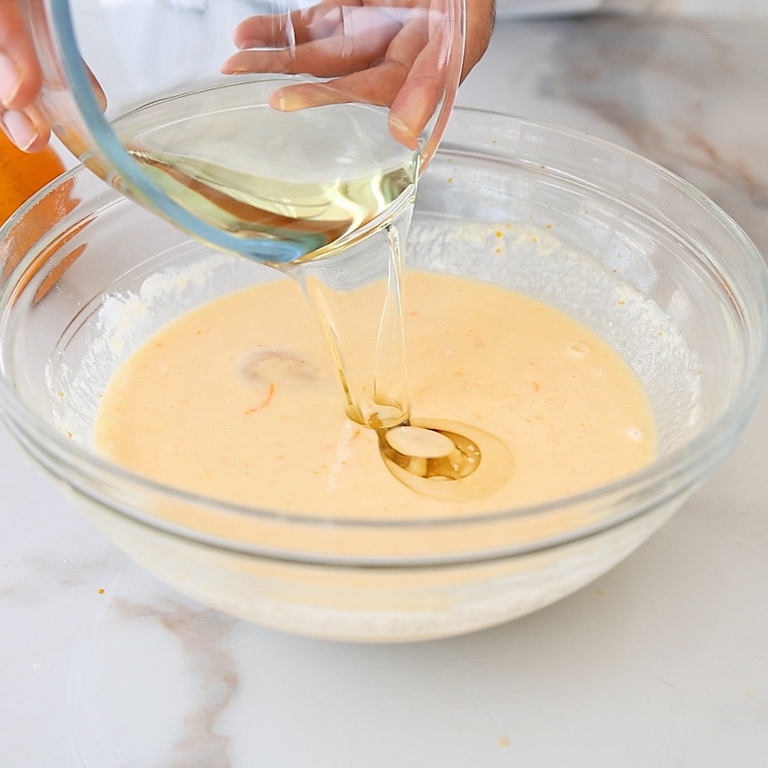 A person is pouring oil from a glass bowl into a larger bowl filled with yellow cake batter on a marble countertop.
