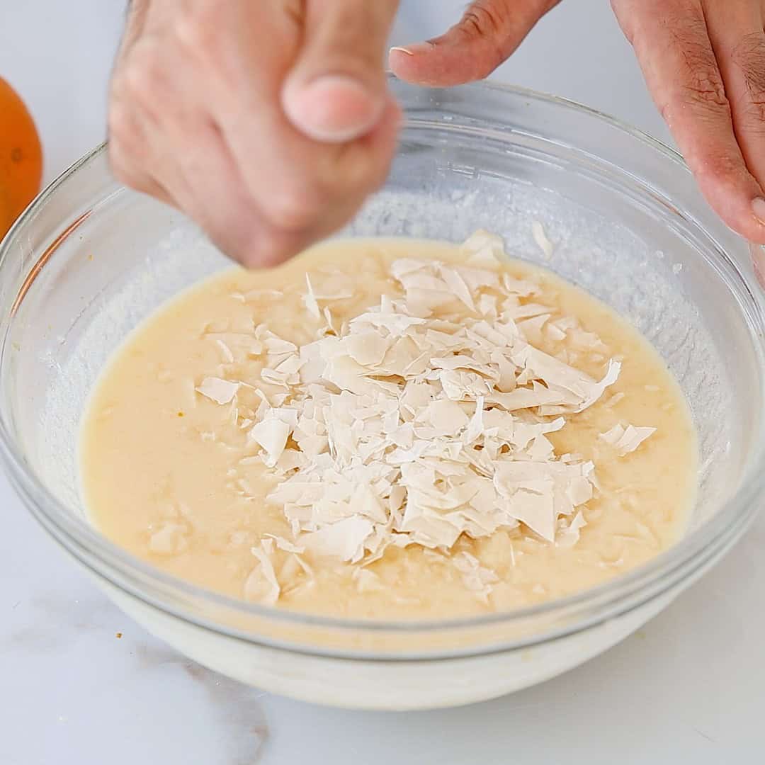 A person adds crumbled phyllo dough to a bowl of batter, preparing to mix the ingredients together by hand. An orange is partially visible on the side of the white countertop.