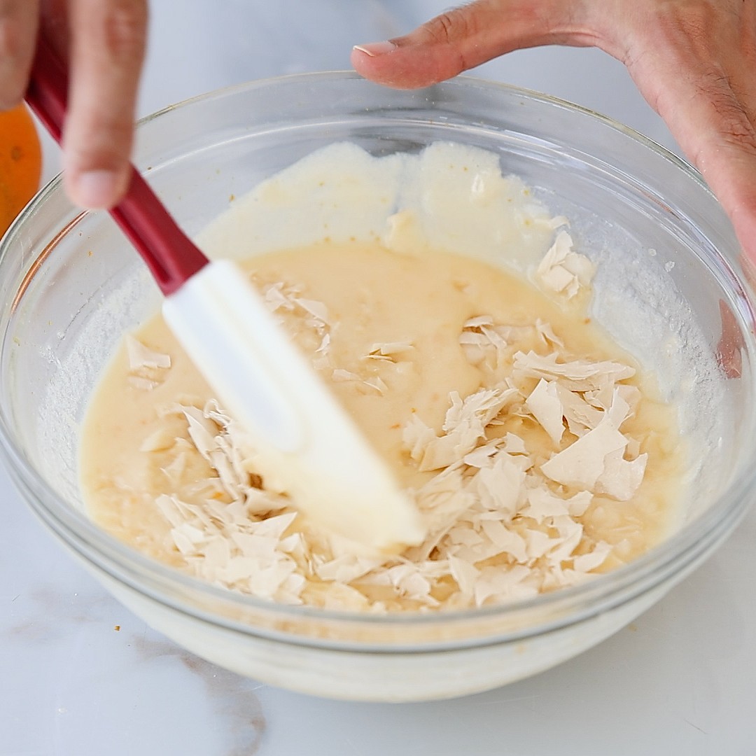 A hand uses a red spatula to mix phyllo dough pieces into a bowl of creamy batter on a white surface.