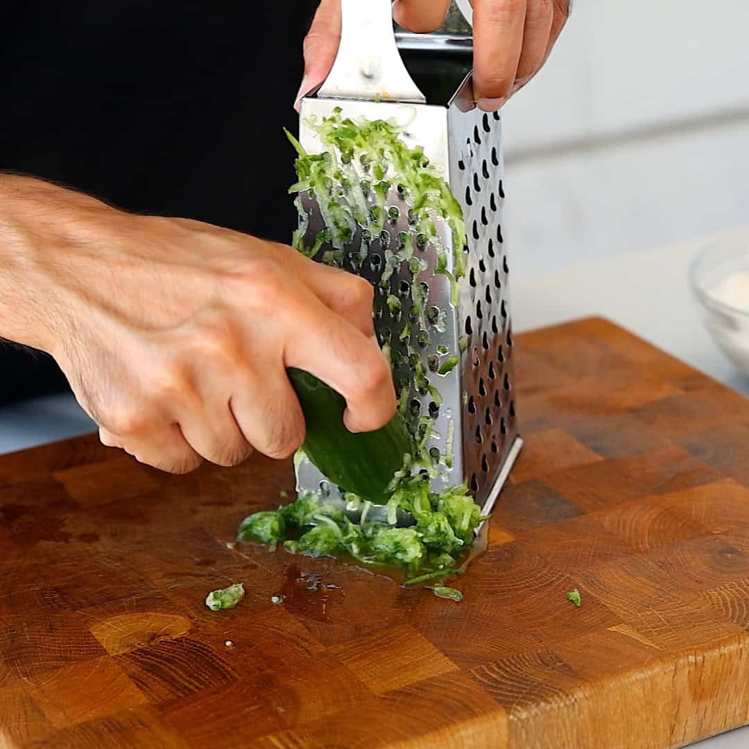 A person grates a cucumber, on a box grater over a wooden cutting board. Grated pieces collect at the base of the grater.