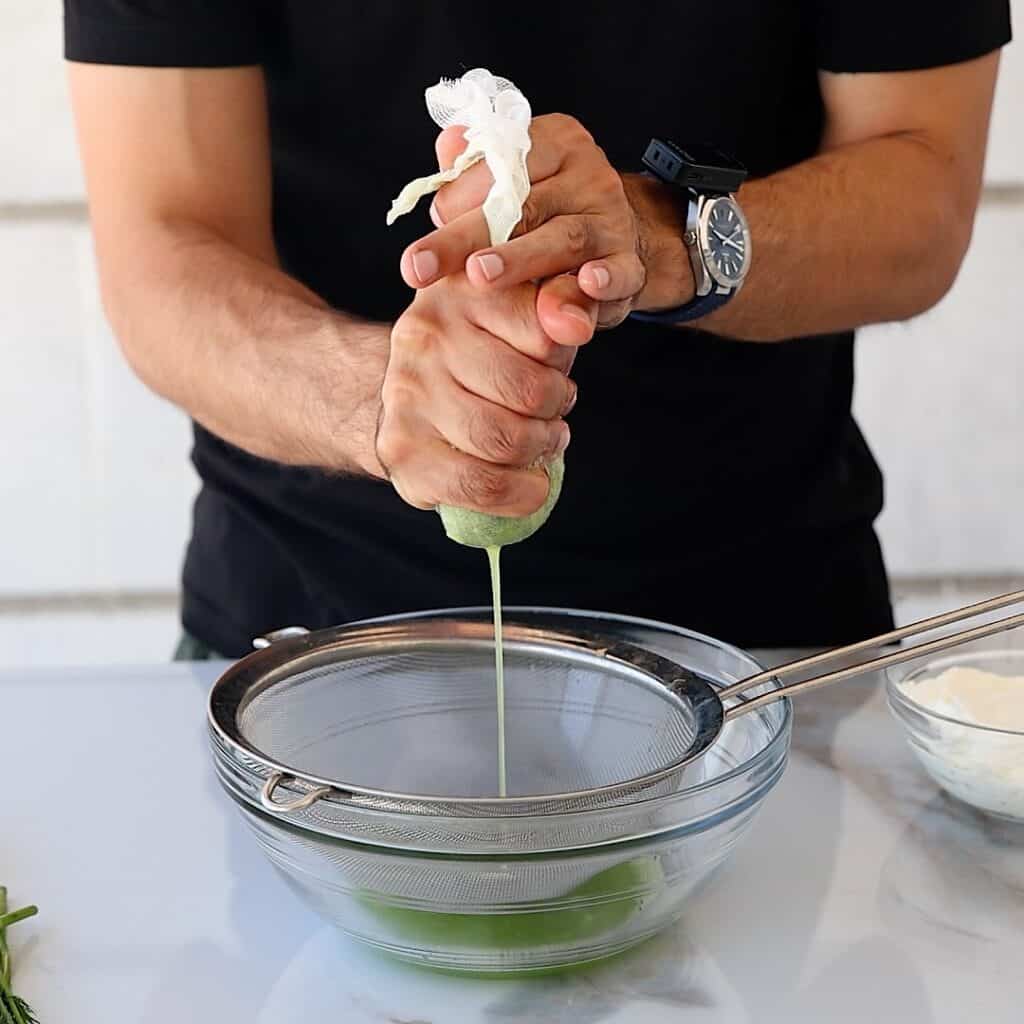 A person squeezes green liquid from a cucumber over a mesh strainer and glass bowl, with a watch on their wrist, preparing ingredients on a countertop.