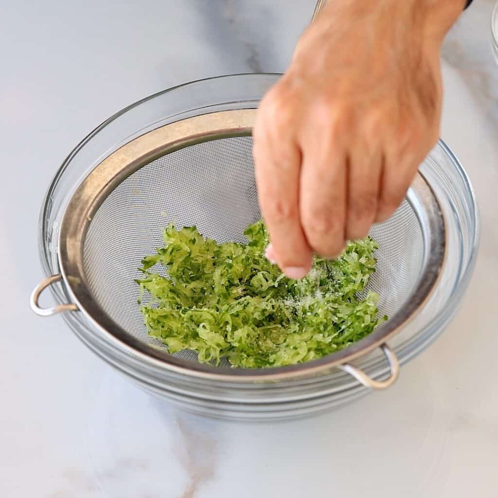A hand sprinkles salt over shredded cucumber i in a metal strainer set above a glass bowl on a light-colored countertop.