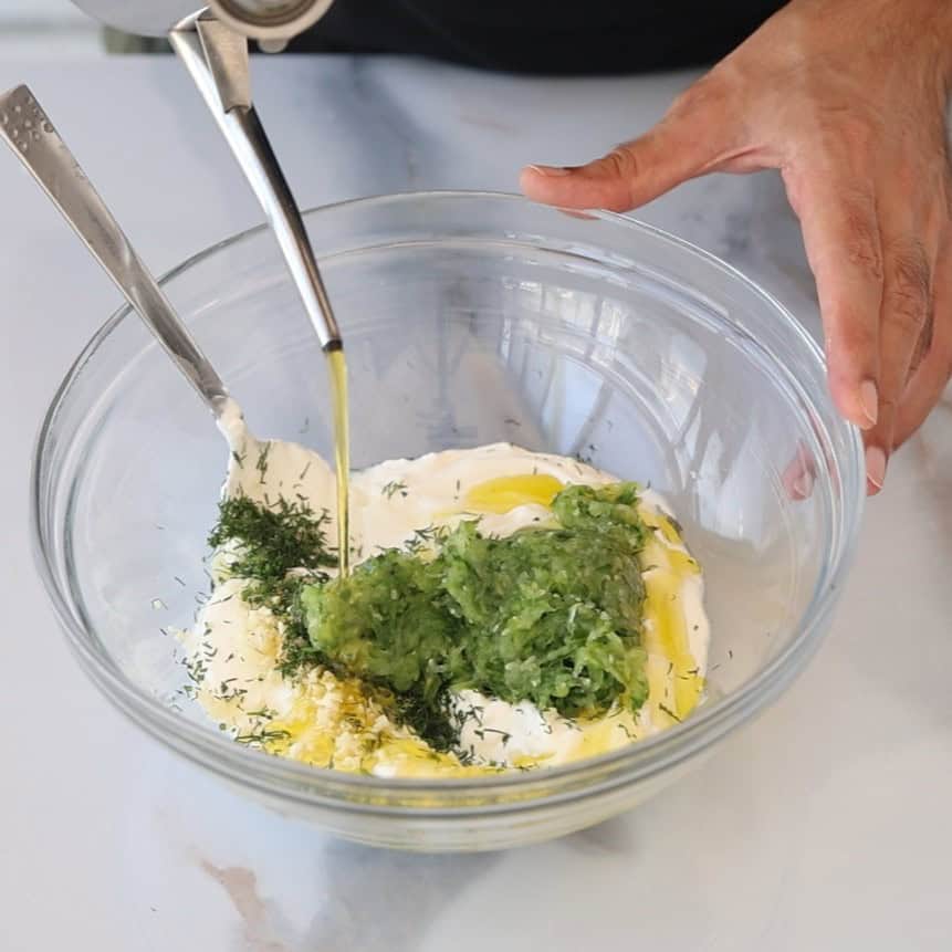 A hand pours olive oil into a glass bowl containing Greek yogurt, grated cucumber, herbs, and garlic, with two spoons resting inside, preparing tzatziki sauce.
