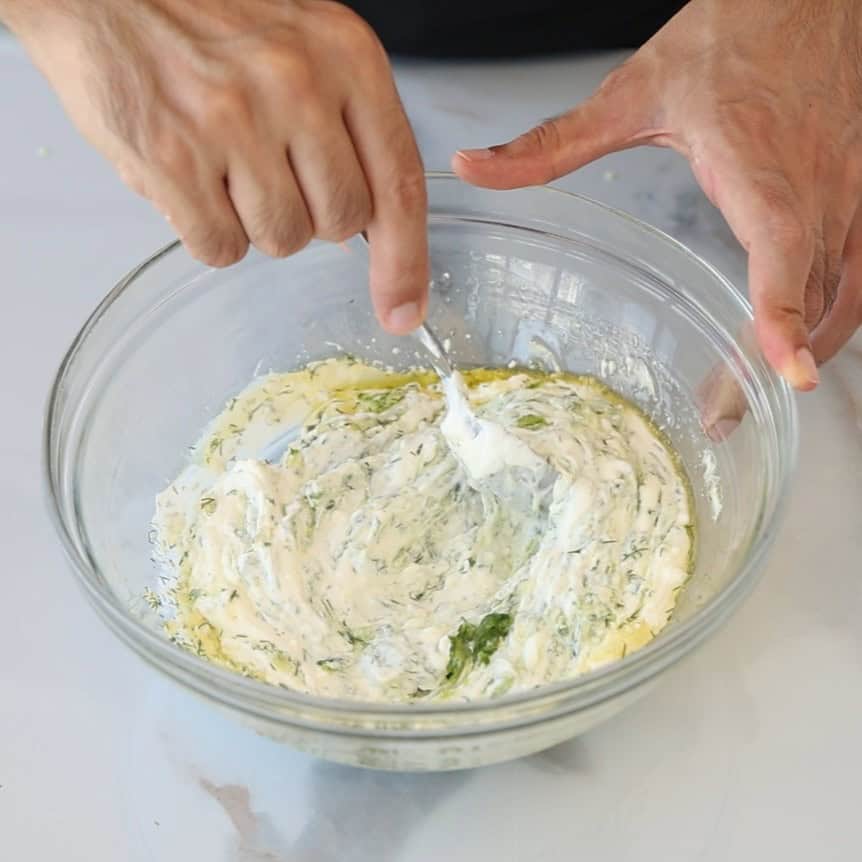 Two hands mix a creamy tzatziki sauce in a clear glass bowl with a spoon on a white surface.