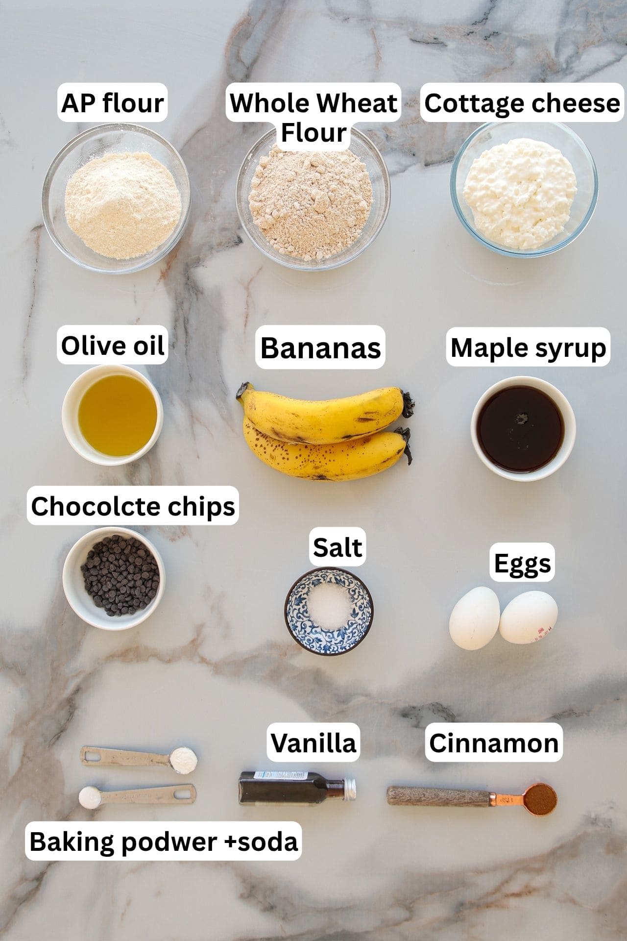 Overhead view of labeled baking ingredients on a marble surface, including flour, whole wheat flour, cottage cheese, olive oil, bananas, maple syrup, chocolate chips, salt, eggs, vanilla, cinnamon, and baking powder with baking soda.