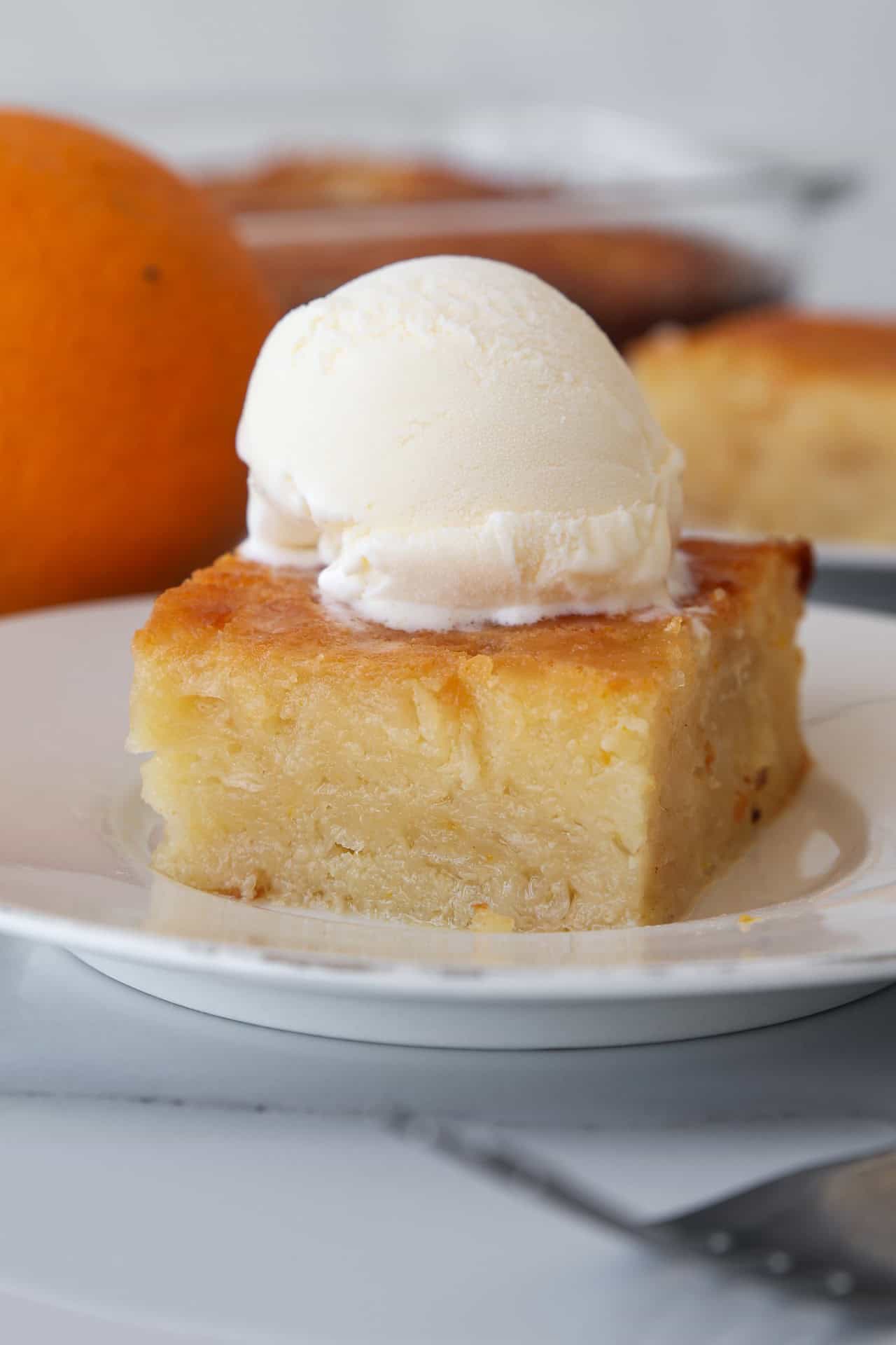 A slice of moist Greek orange cake (also known as portokalopita)  topped with a scoop of vanilla ice cream sits on a white plate, with an orange and more cake in the background.