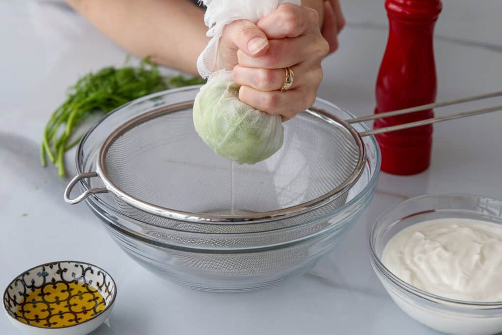 A hand squeezes liquid from grated cucumber in cheesecloth over a strainer and glass bowl, with dill, pepper mill, olive oil, and yogurt on a white countertop.