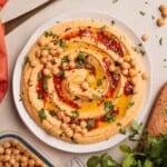 Plate of hummus with harissa dup topped with whole chickpeas, olive oil, red spices, and chopped parsley, surrounded by fresh cilantro and a container of chickpeas on a light surface.