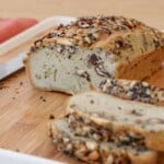 A loaf of tahini-seeded bread with visible nuts, partially sliced on a wooden cutting board with a knife and a red cloth in the background.