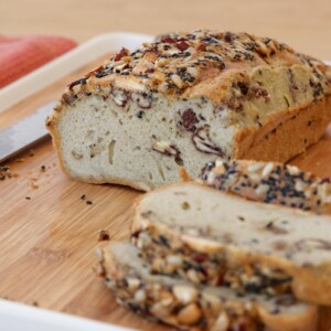 A loaf of tahini-seeded bread with visible nuts, partially sliced on a wooden cutting board with a knife and a red cloth in the background.