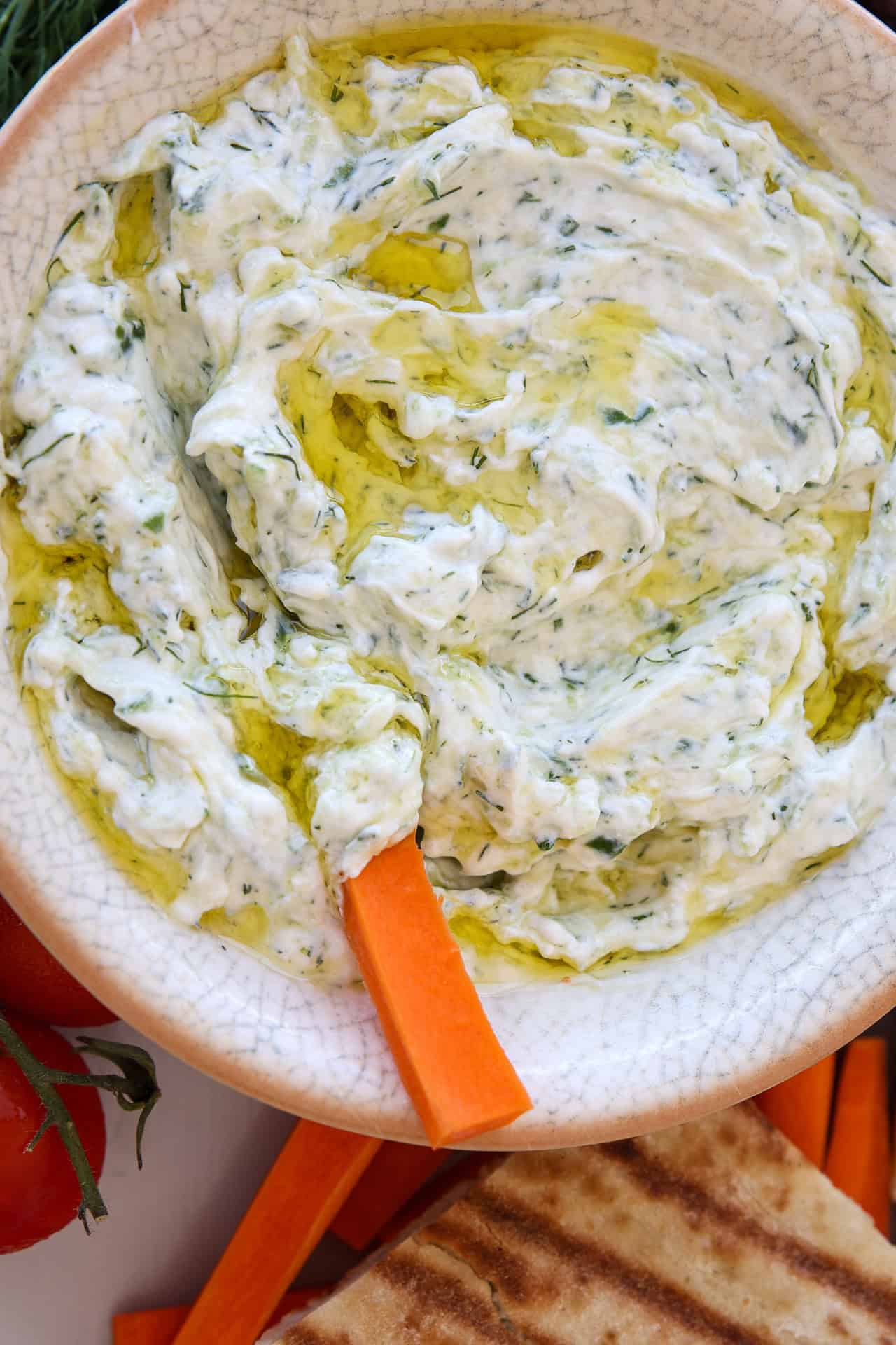 A bowl of thick Greek tzatziki dip with dill and olive oil, served with carrot sticks and pita bread.