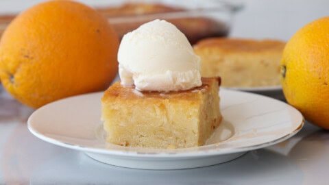 Authentic Portokalopita Recipe (Greek Orange Cake) A slice of orange cake topped with a scoop of vanilla ice cream sits on a white plate, with whole oranges and more cake visible in the background.