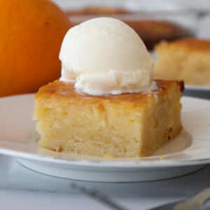 A square piece of moist Greek yogurt and orange cake topped with a scoop of vanilla ice cream sits on a white plate, with an orange in the background.