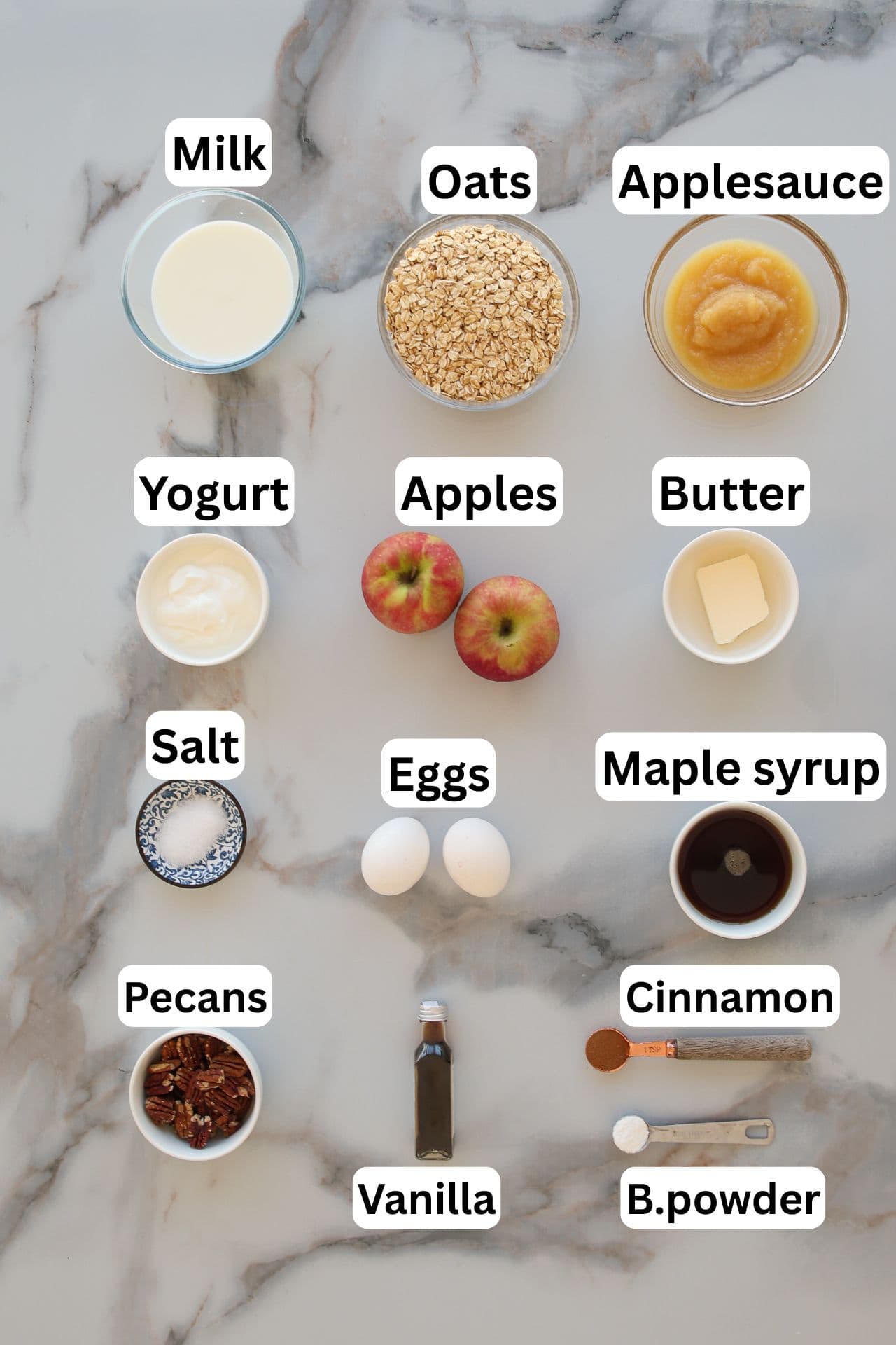Ingredients for baked apple oatmeal on a marble surface: milk, oats, applesauce, yogurt, apples, butter, salt, eggs, maple syrup, pecans, vanilla, cinnamon, and baking powder, each labeled with text.