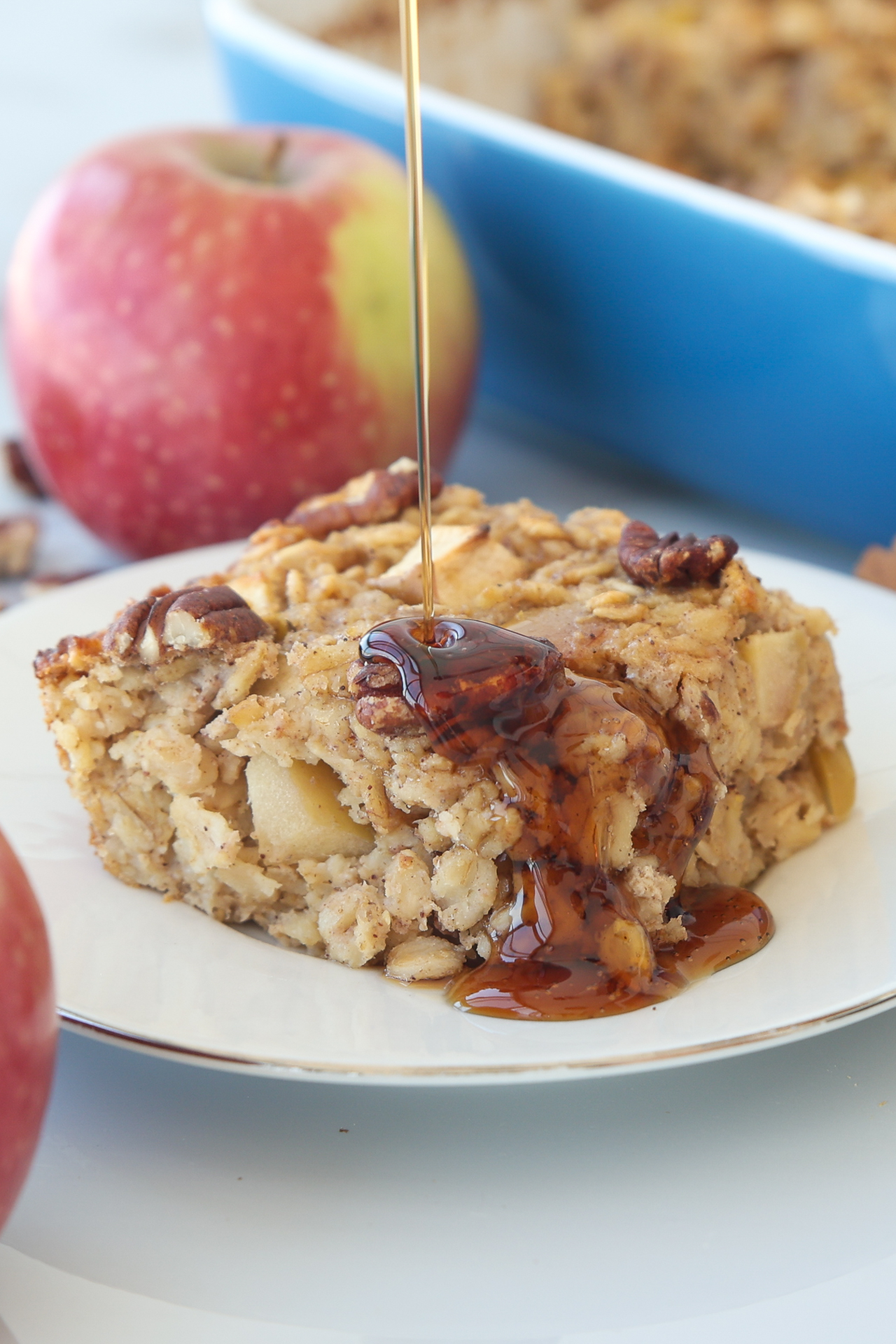 A slice of baked oatmeal with apple chunks and pecans sits on a plate. Syrup is being poured on top, and a red apple and blue baking dish are visible in the background.