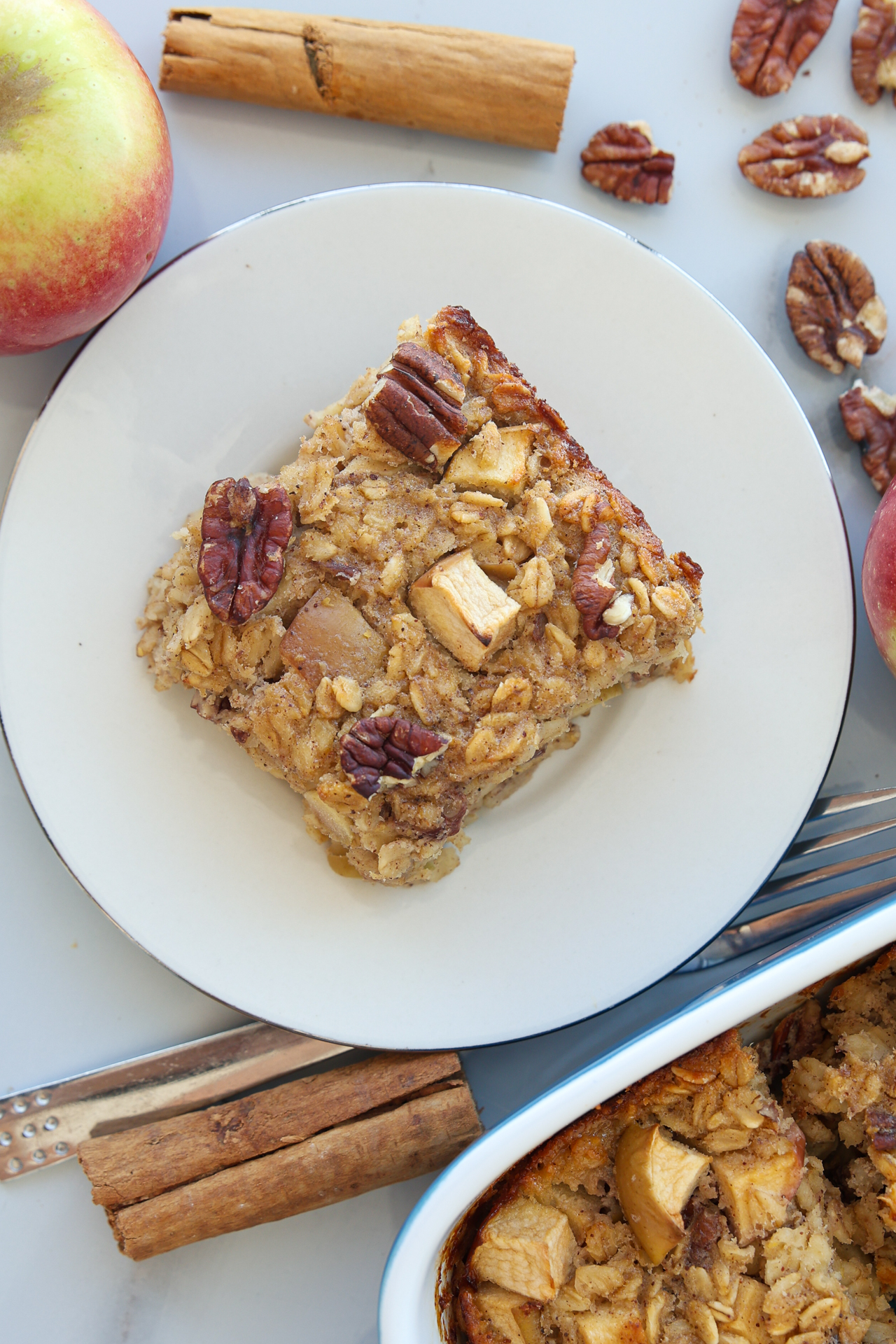 A square slice of healthy apple baked oatmeal with pecans on a white plate, surrounded by apples, cinnamon sticks, and a baking dish on a light surface.