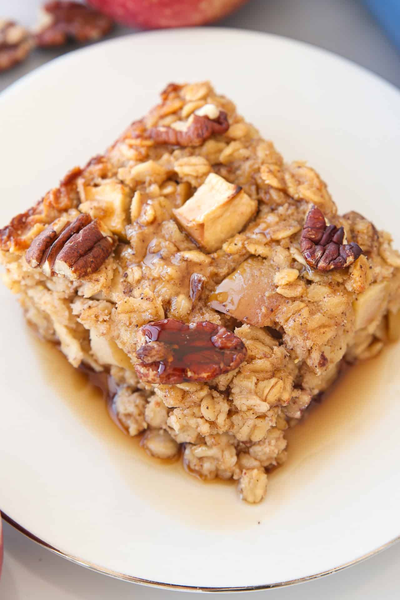A square slice of baked apple pie oatmeal topped with pecans and apple chunks sits on a white plate, drizzled with syrup.