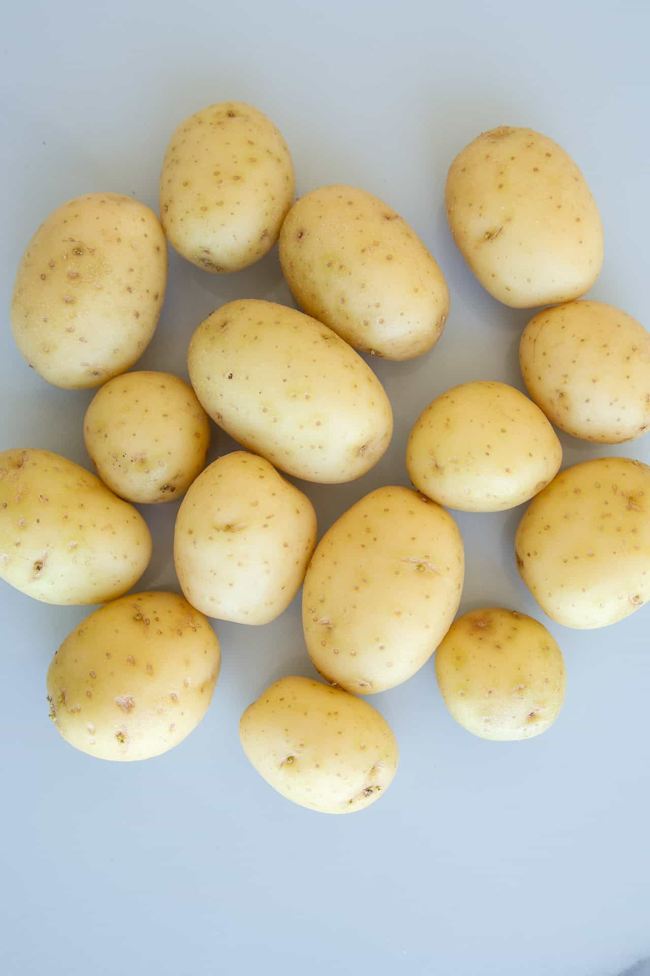 Fifteen small, unpeeled, light brown potatoes are arranged on a flat, light gray surface.