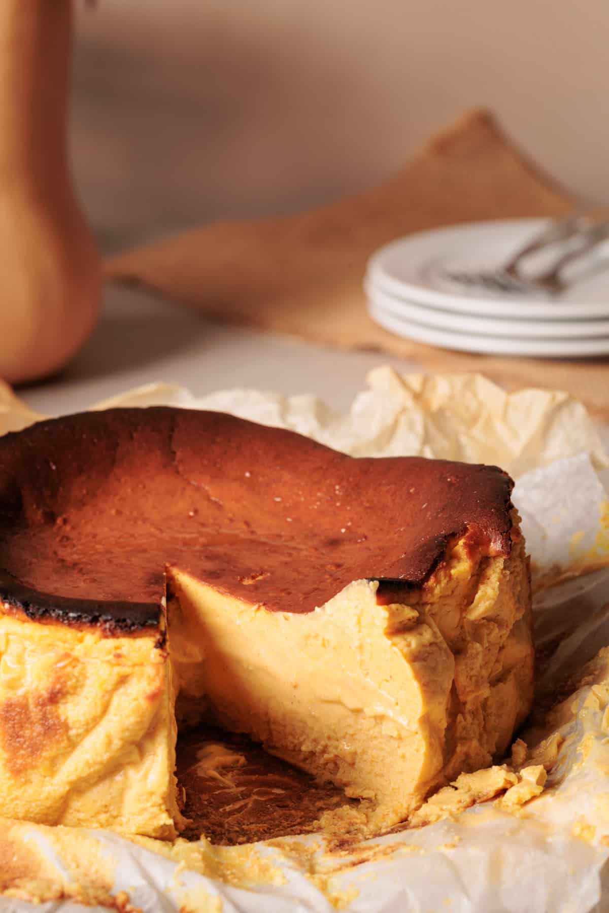 A creamy, golden-brown pumpkin baqsue cheesecake sits on parchment paper with a large slice removed, revealing its dense, rich interior. Stacked white plates and a blurred background are visible.
