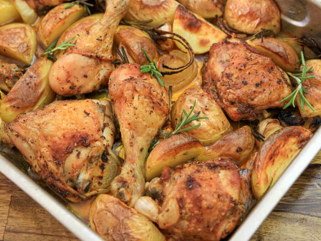 Roasted chicken drumsticks and thighs with golden potatoes, onions, and sprigs of rosemary in a baking tray.