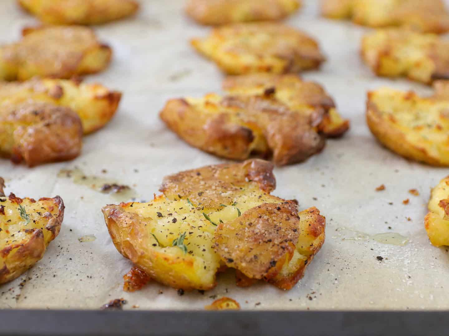 Crispy smashed potatoes seasoned with herbs and cracked black pepper are baked on a parchment-lined tray.