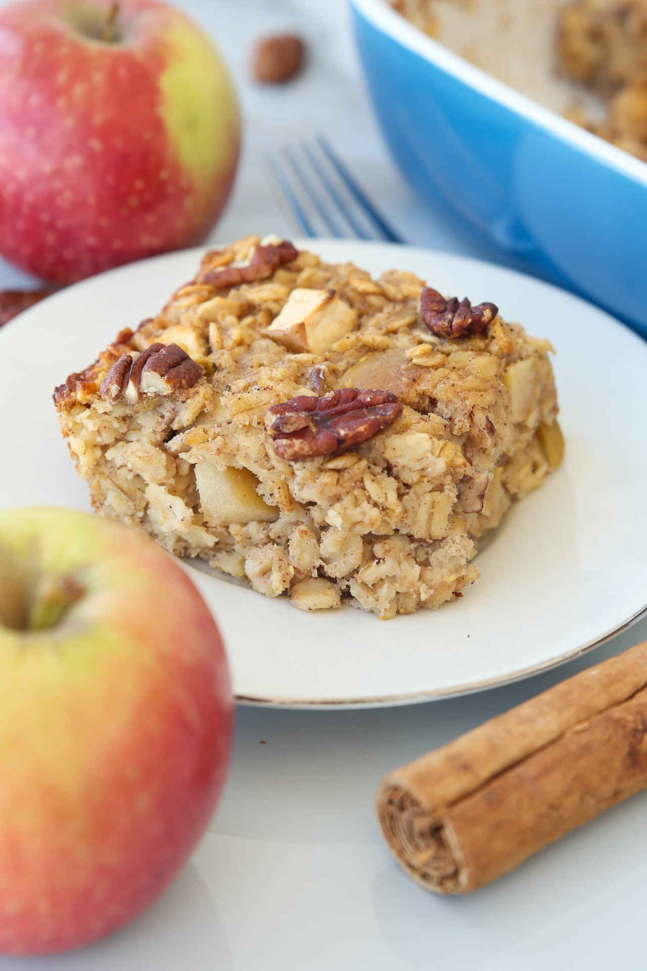 A square slice of Baked Apple Oatmeal topped with pecans sits on a white plate, surrounded by fresh apples, a cinnamon stick, and a blue baking dish in the background.