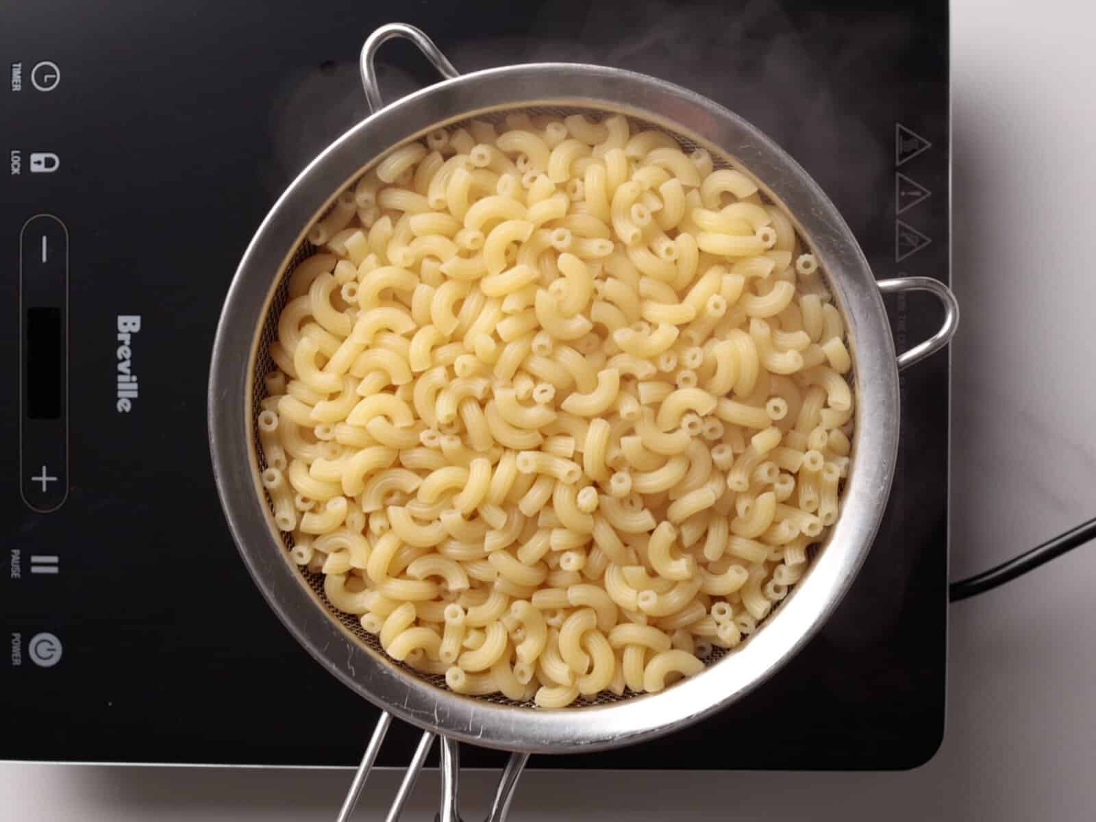 A metal strainer filled with cooked elbow macaroni pasta rests on top of a pot or electric stovetop, with steam rising from the hot pasta.