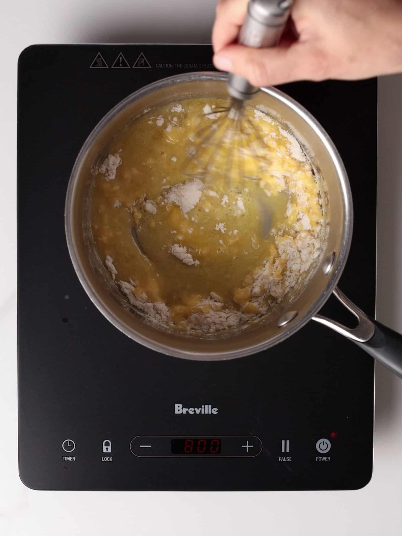 A hand whisks flour and melted butter in a saucepan on an electric cooktop, starting to make a roux.