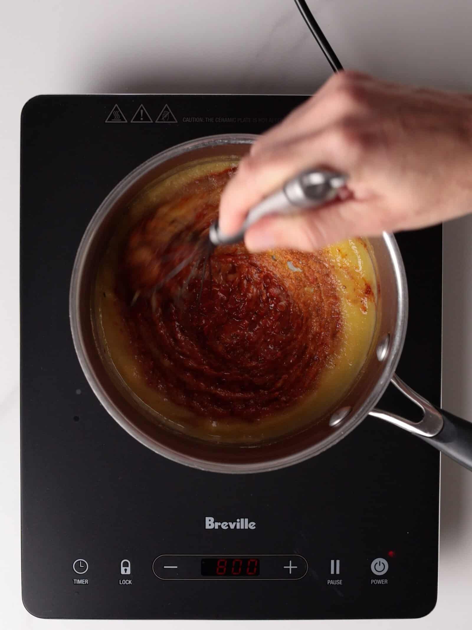 A hand uses a whisk to mix a harissa-based roux for a spicy mac and cheese.