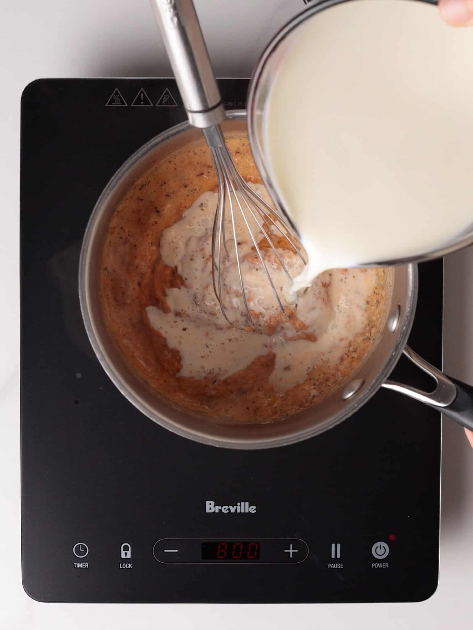 A saucepan on an induction cooktop contains a whisked roux, while milk is poured in from a glass measuring cup.