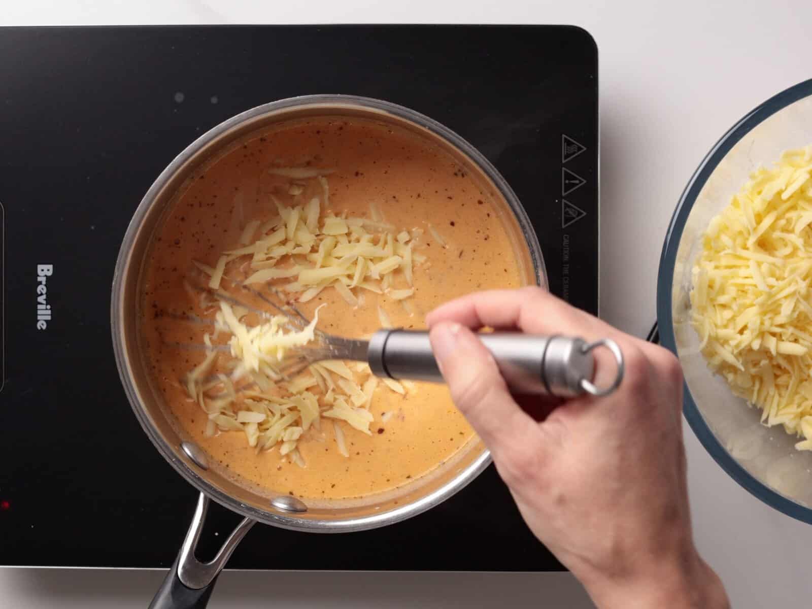 A hand stirs shredded cheese into a creamy orange sauce in a saucepan on a stovetop, with a bowl of more shredded cheese nearby.