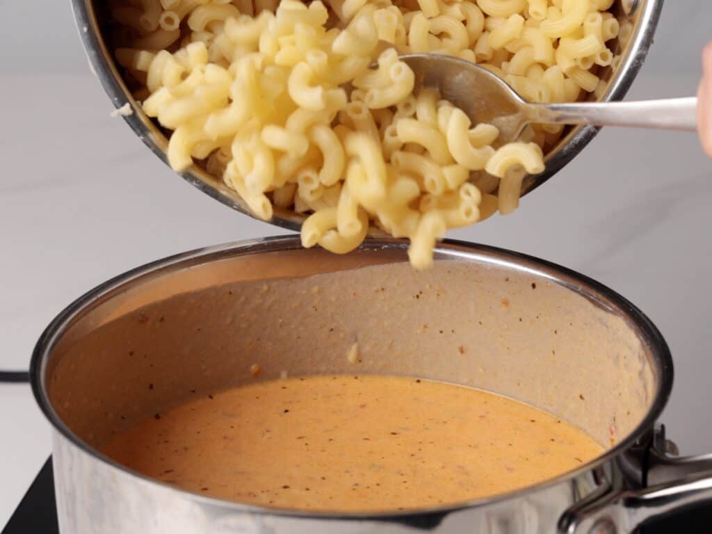 Cooked macaroni being spooned from a pot into a saucepan filled with creamy cheese sauce, ready to be mixed for macaroni and cheese.