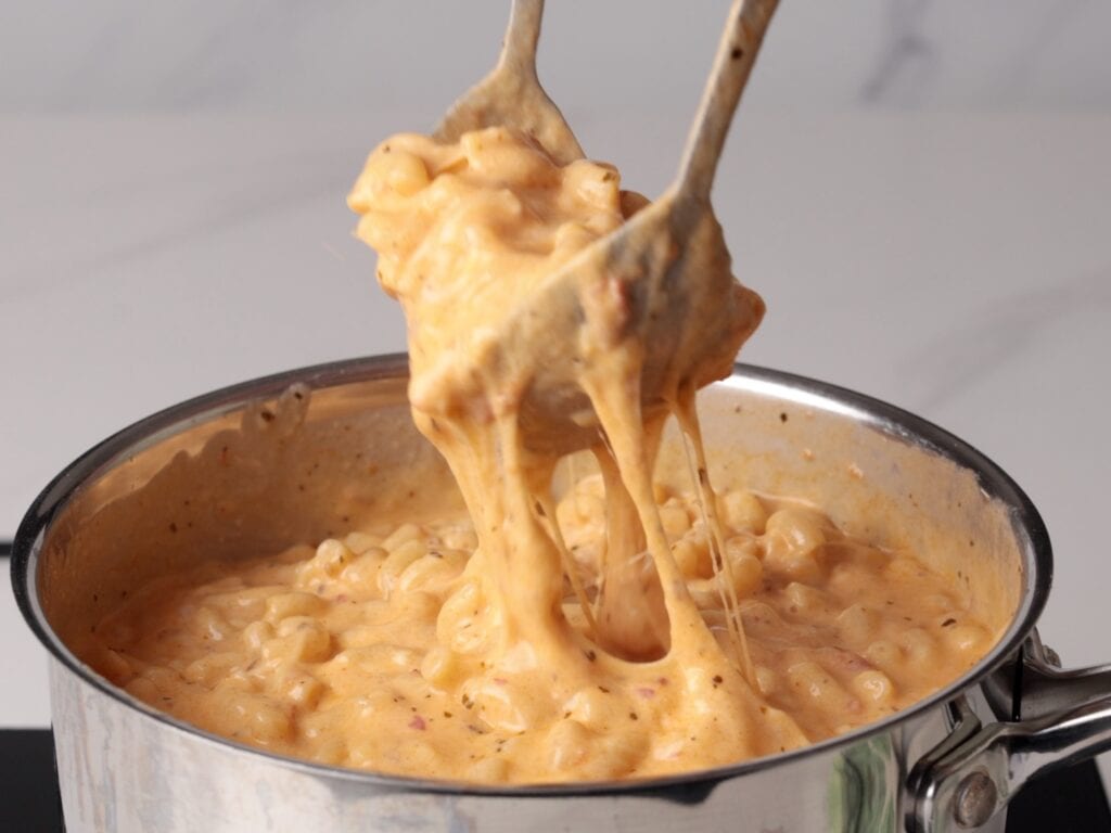 A pot of creamy harissa macaroni and cheese with stretchy, melted cheese being lifted by two utensils.