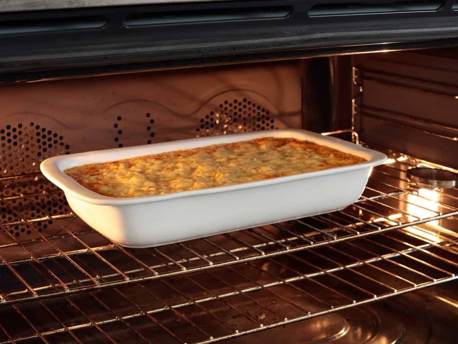 A rectangular white dish with a baked spicy mac and cheese sits on an oven rack, with a golden, bubbly cheese topping visible under the oven light.