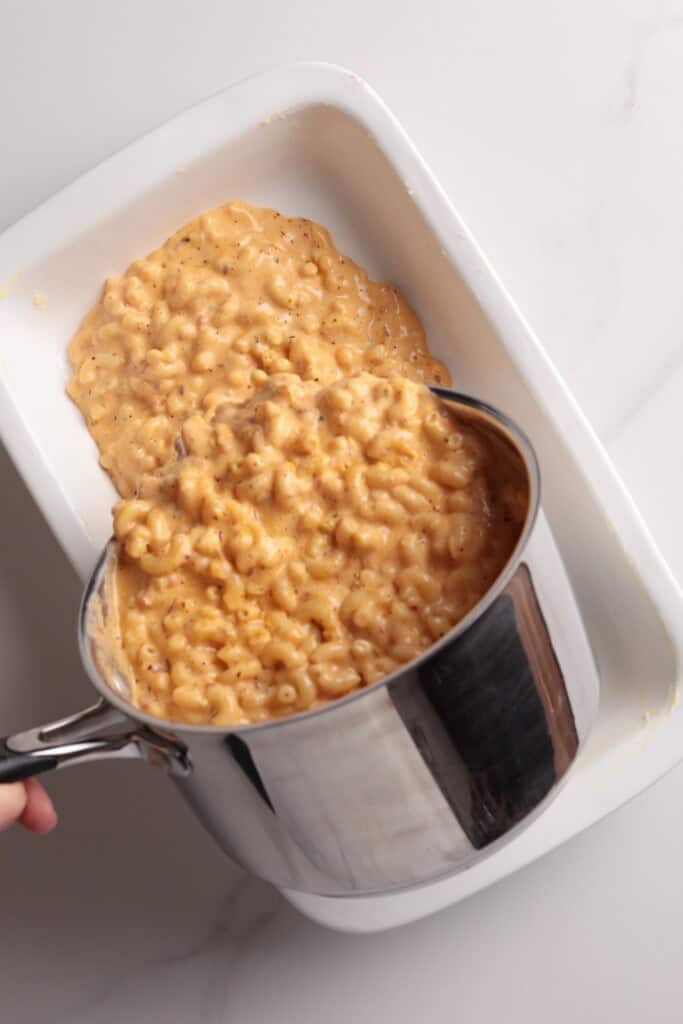 A pot of creamy macaroni and cheese is being poured into a white rectangular baking dish on a white surface.