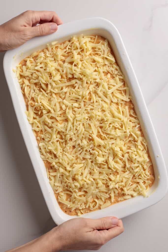 Hands hold a white baking dish filled with an unbaked casserole topped with shredded cheese on a white surface.