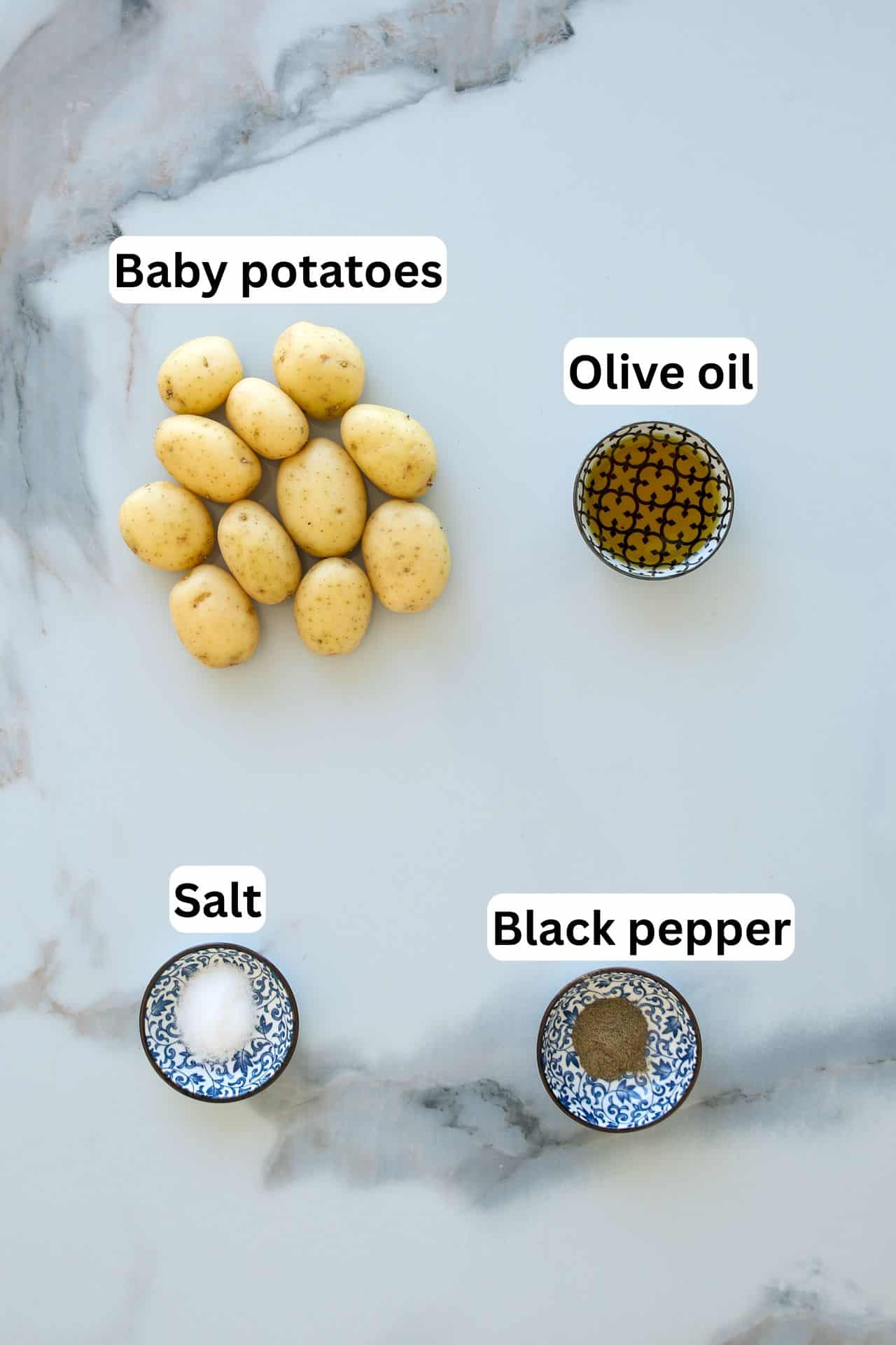 Four labeled ingredients on a marble surface: a pile of baby potatoes, a small bowl of olive oil, a small bowl of salt, and a small bowl of black pepper.