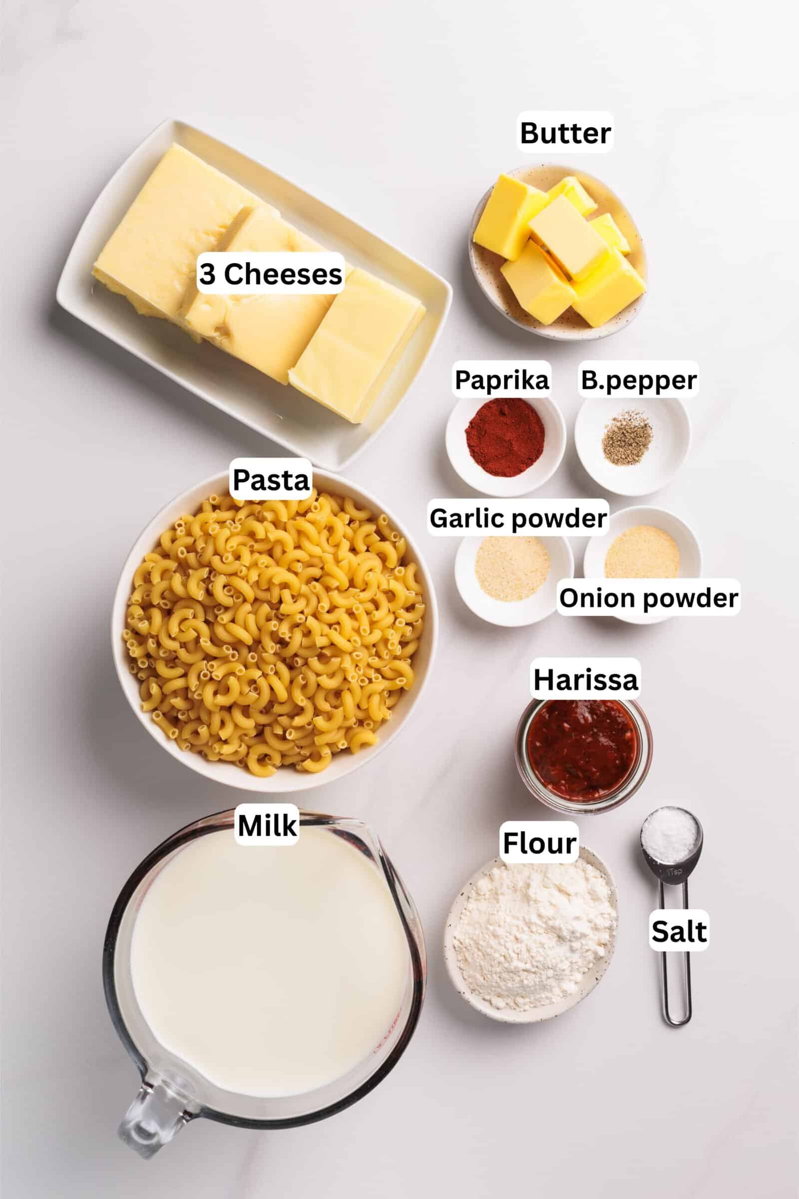 Top view of labeled ingredients for spicy macaroni and cheese: pasta, milk, flour, salt, harissa, onion powder, garlic powder, black pepper, paprika, butter, and three types of cheese on a white surface.
