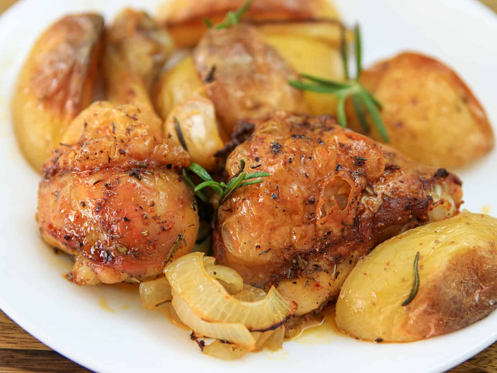 Sheet pan roasted chicken and golden potatoes, onions, and sprigs of rosemary on a white plate. The chicken skin is crispy and browned, and the potatoes look tender and seasoned.