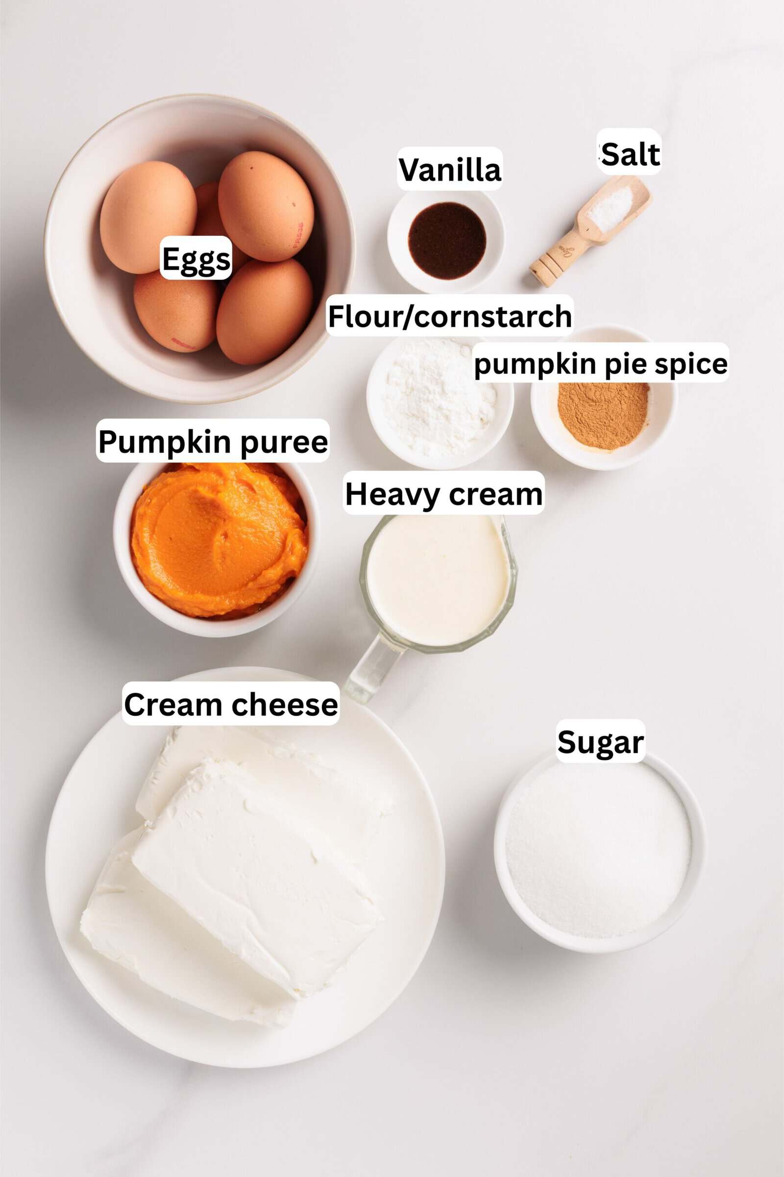 Bowls and plates containing eggs, vanilla, salt, flour or cornstarch, pumpkin pie spice, pumpkin puree, heavy cream, cream cheese, and sugar, all labeled, are arranged on a white surface.