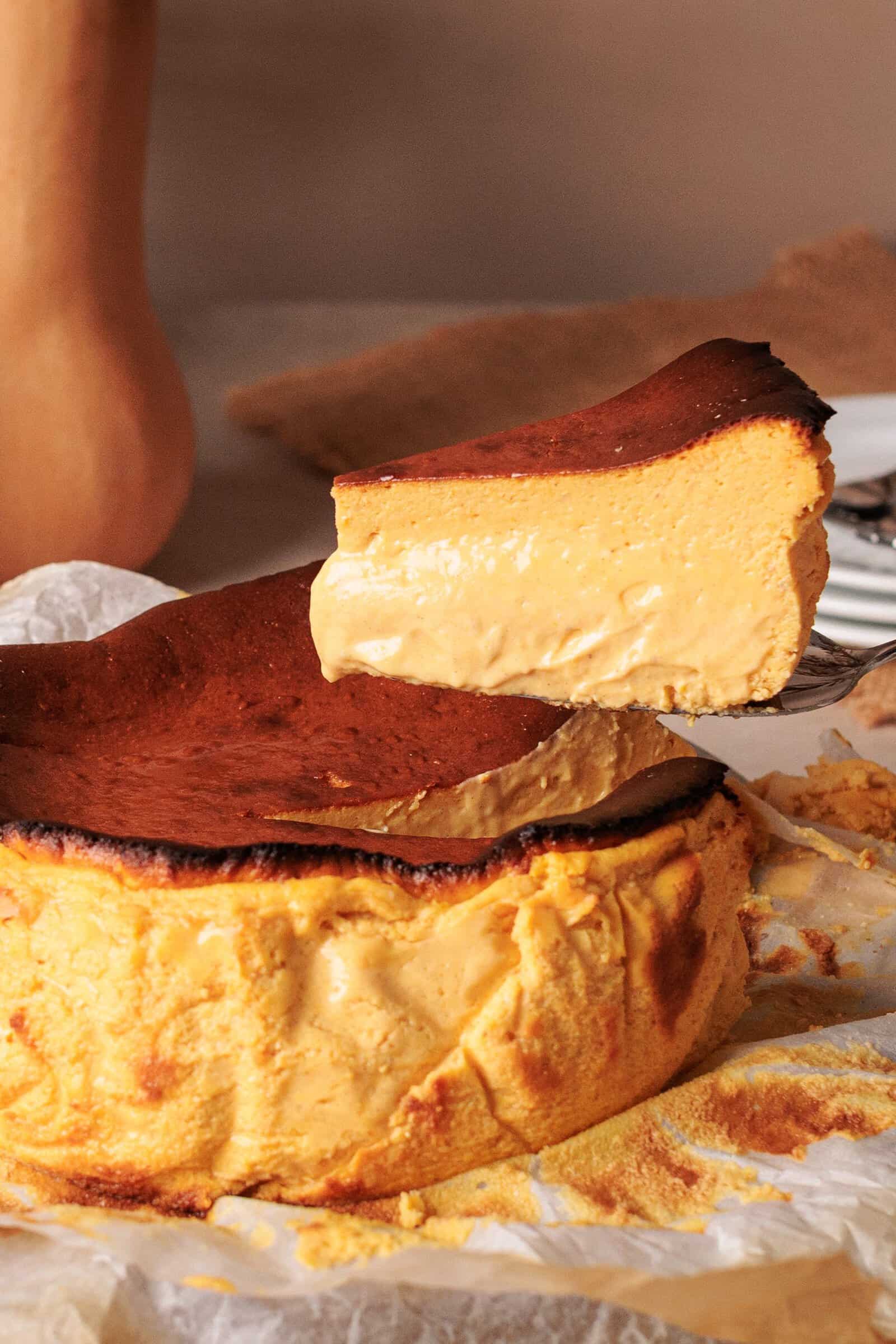 A slice of creamy pumpkin Basque cheesecake is being lifted from the whole cake, which sits on parchment paper. The cheesecake has a caramelized top and a smooth, dense texture.