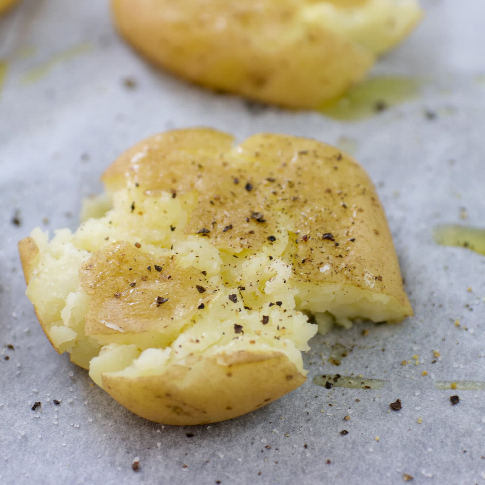 A cooked potato smashed flat, topped with black pepper and seasoning, sits on parchment paper with a drizzle of oil.