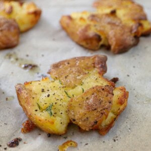 Close-up of smashed roasted potatoes on parchment paper, seasoned with black pepper and herbs, with a crispy golden exterior and soft inside.