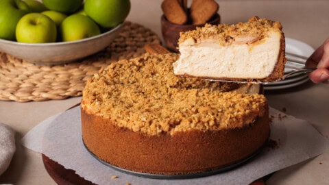 Apple Crumble Cheesecake A hand lifts a slice of apple crumble cheesecake from a whole cake on parchment paper; a bowl of green apples and cookies are in the background.