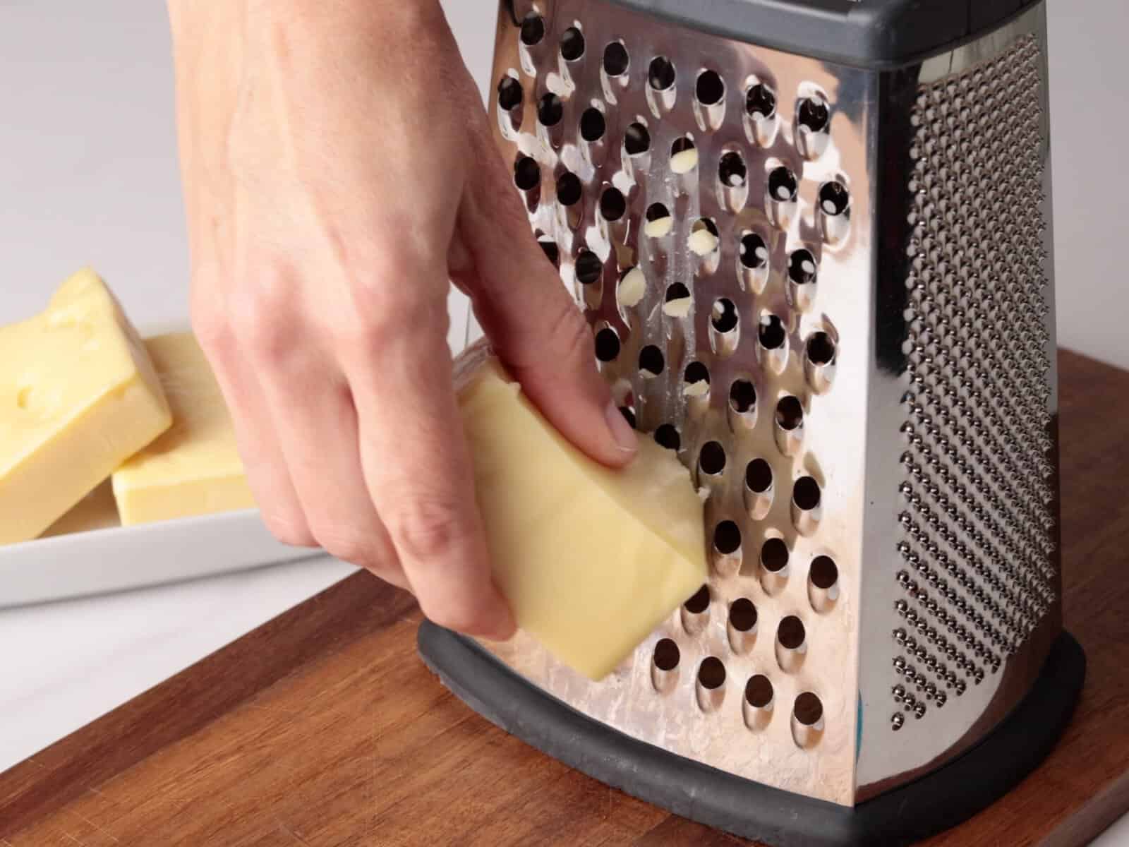 A hand grates a block of cheese on a metal box grater over a wooden surface, with more cheese blocks on a white plate in the background.