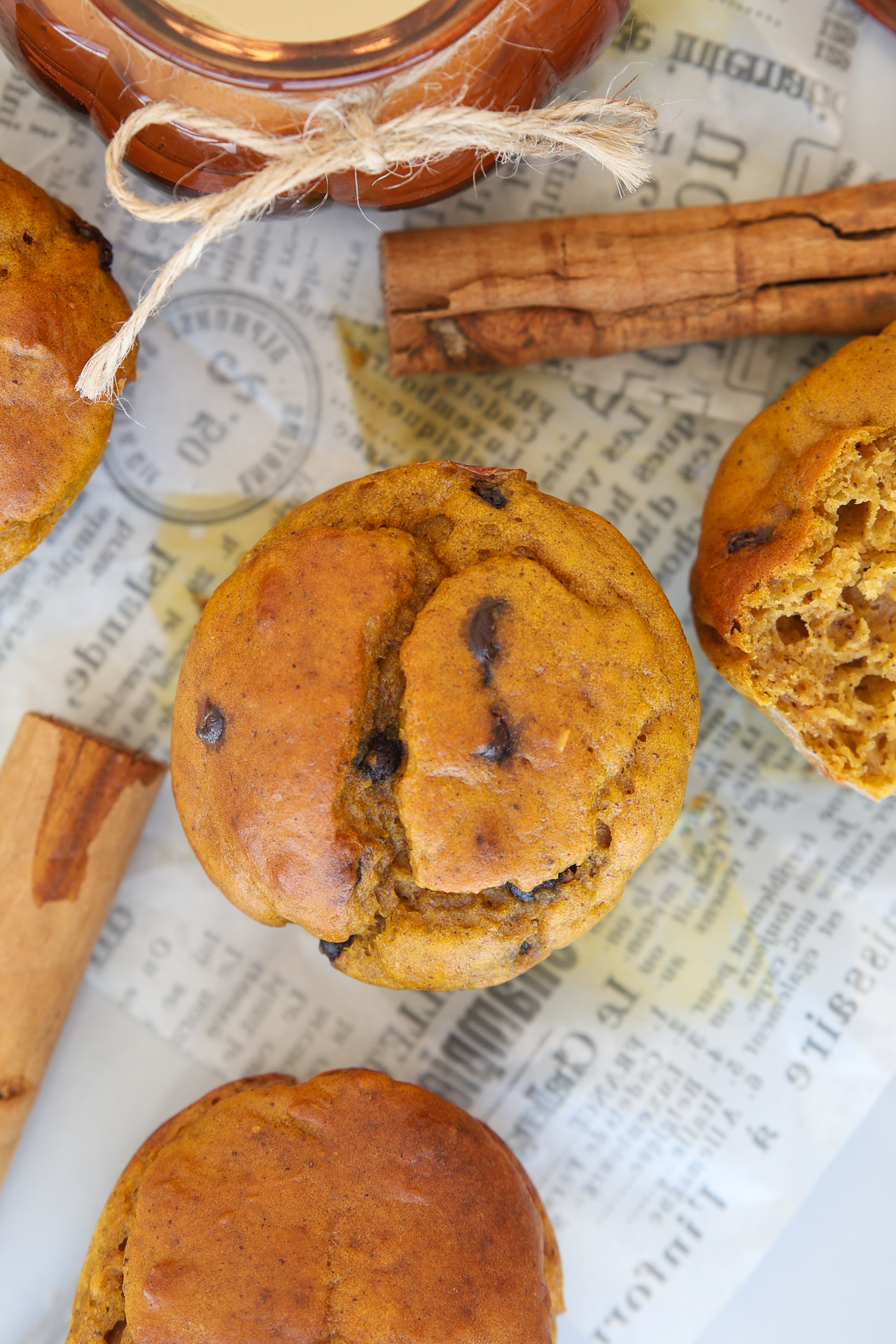 Cottage cheese pumpkin muffins with chocolate chips on newspaper, surrounded by cinnamon sticks and a jar with twine. One muffin is partially broken to reveal a moist inside.