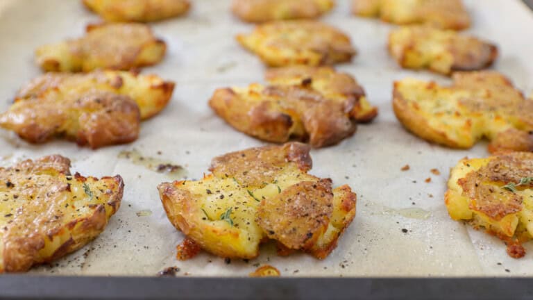 Crispy smashed potatoes on a baking sheet, seasoned with herbs, black pepper, and sea salt, resting on parchment paper after roasting.