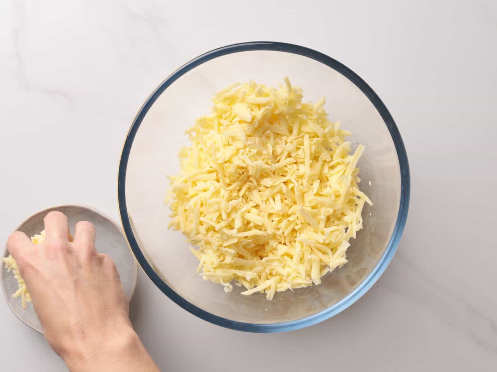 A hand holds a small dish, adding shredded cheese to a clear glass bowl filled with more shredded cheese on a light countertop.
